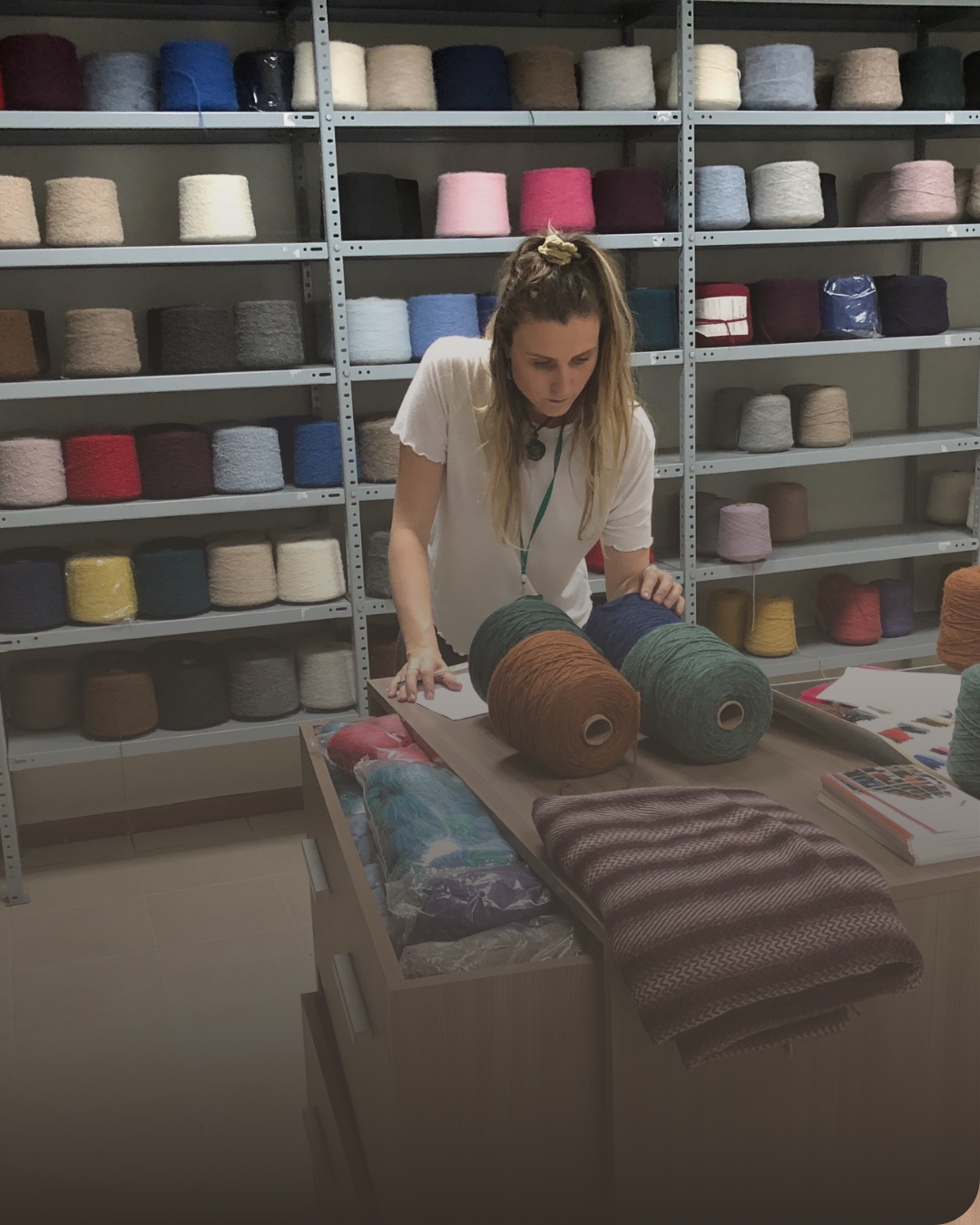 Woman examining large yarn cones in a textile studio with shelves of colorful yarn