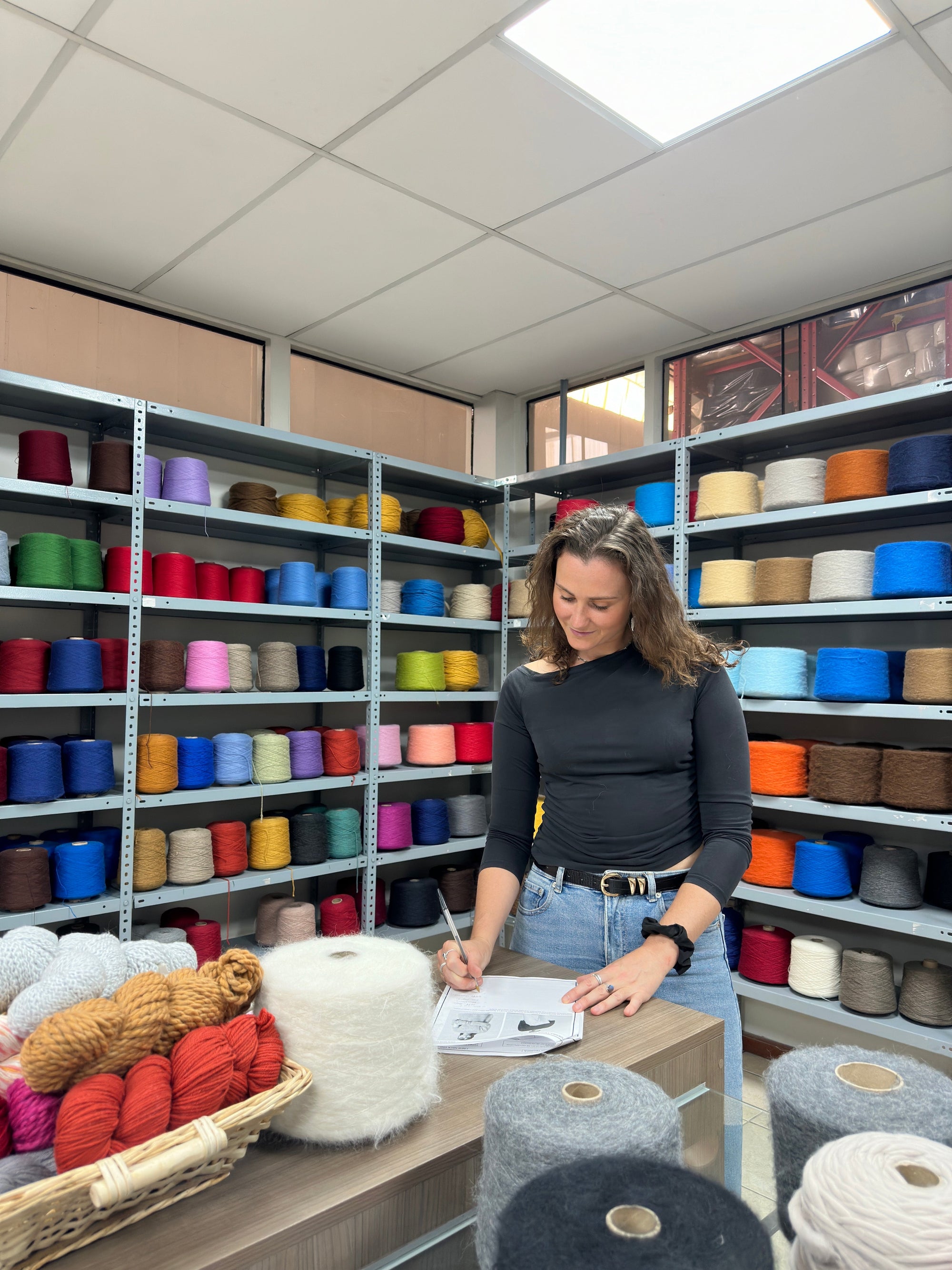 Woman taking notes in yarn shop surrounded by colorful alpaca wool skeins and spools.