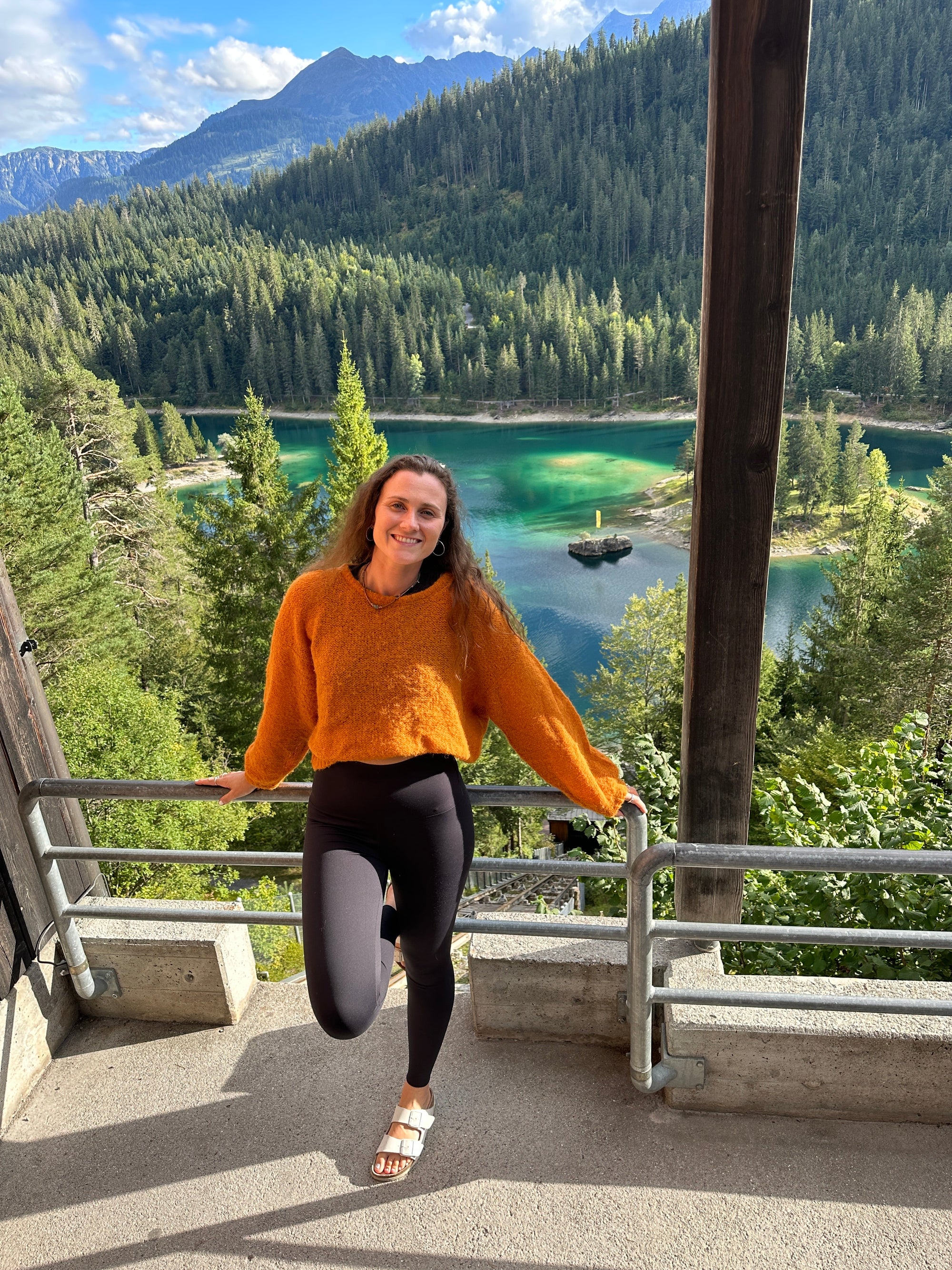 Woman in orange sweater and black leggings posing by scenic mountain lake view