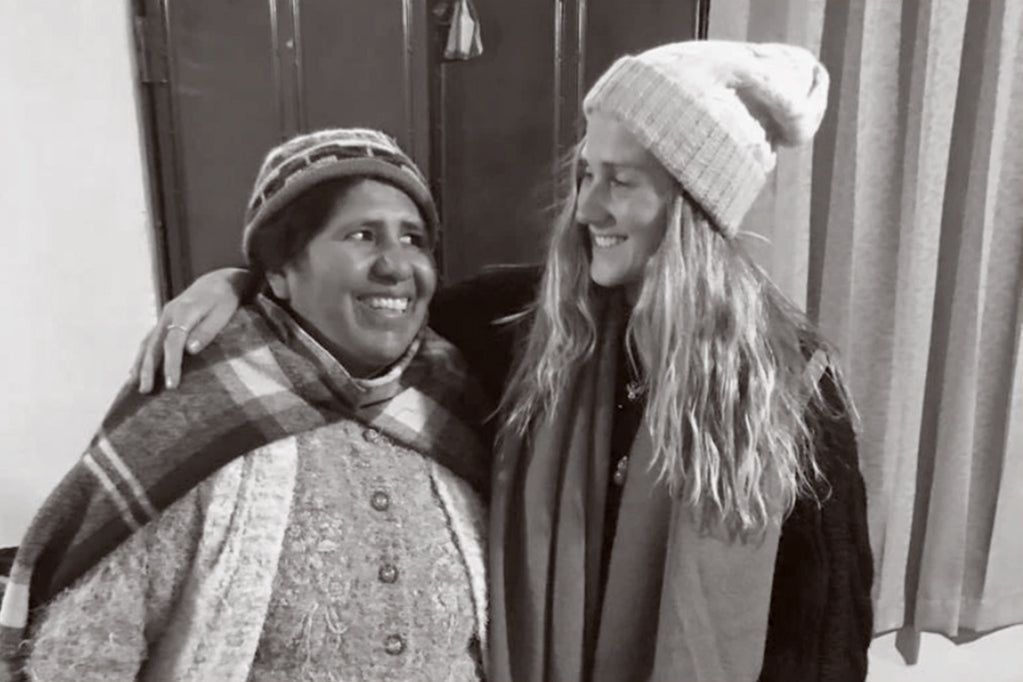 Two women smiling in warm knitwear and scarves, embracing, cozy indoor setting