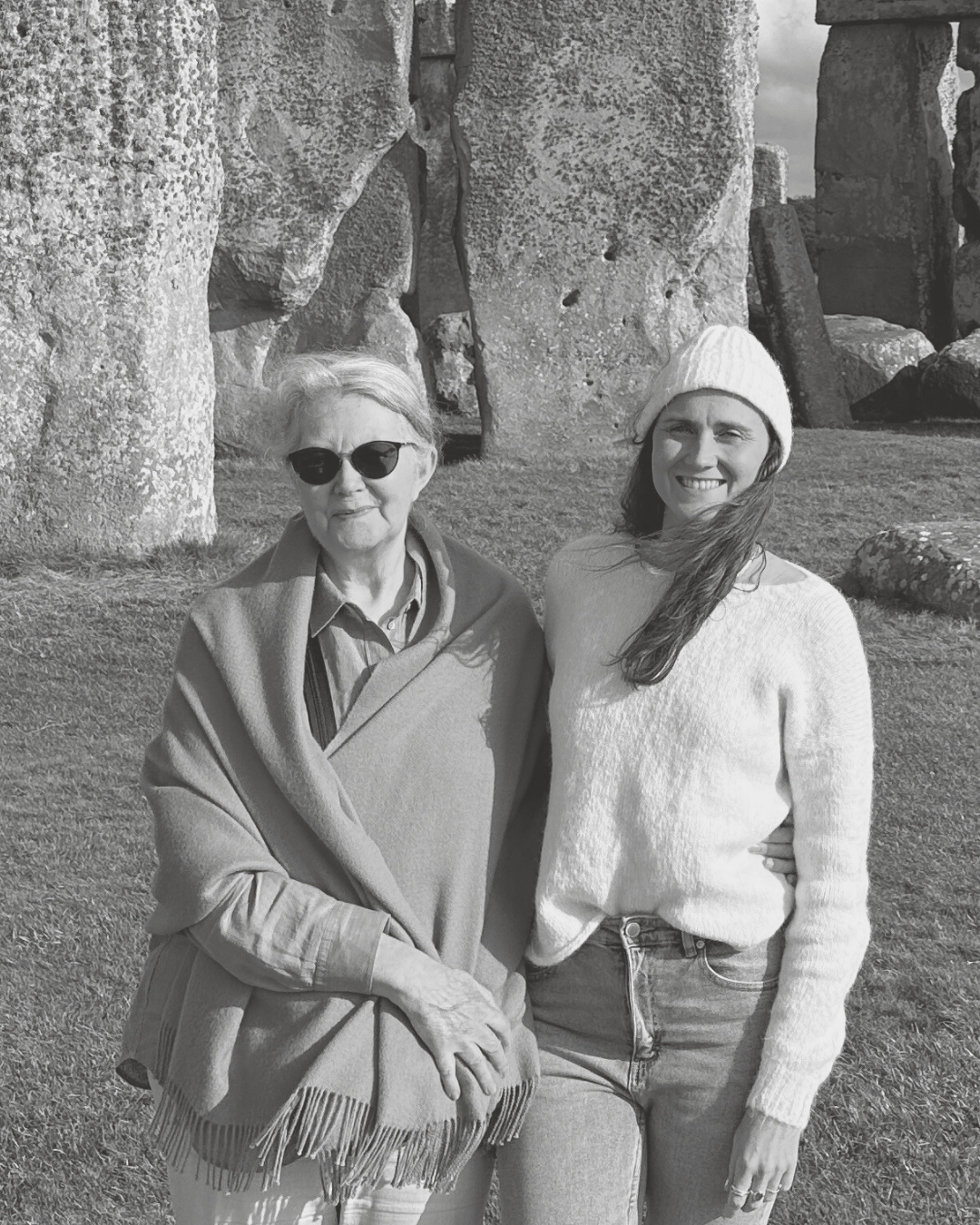 Two women standing in front of large stone ruins on grass, wearing cozy knitwear
