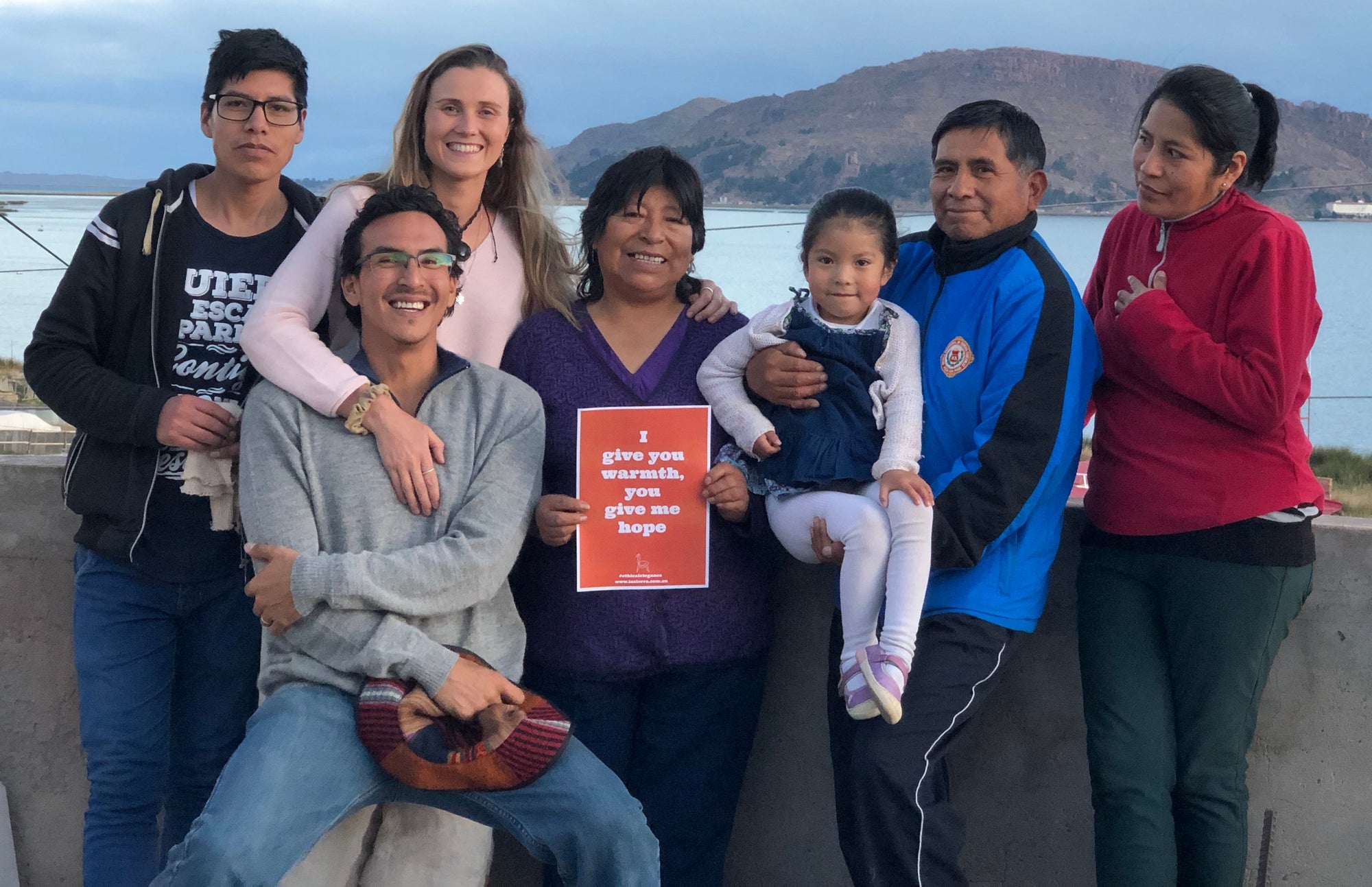 Smiling group of people outdoors by a lake, one holding a sign that says 'I give you warmth, you give me hope'