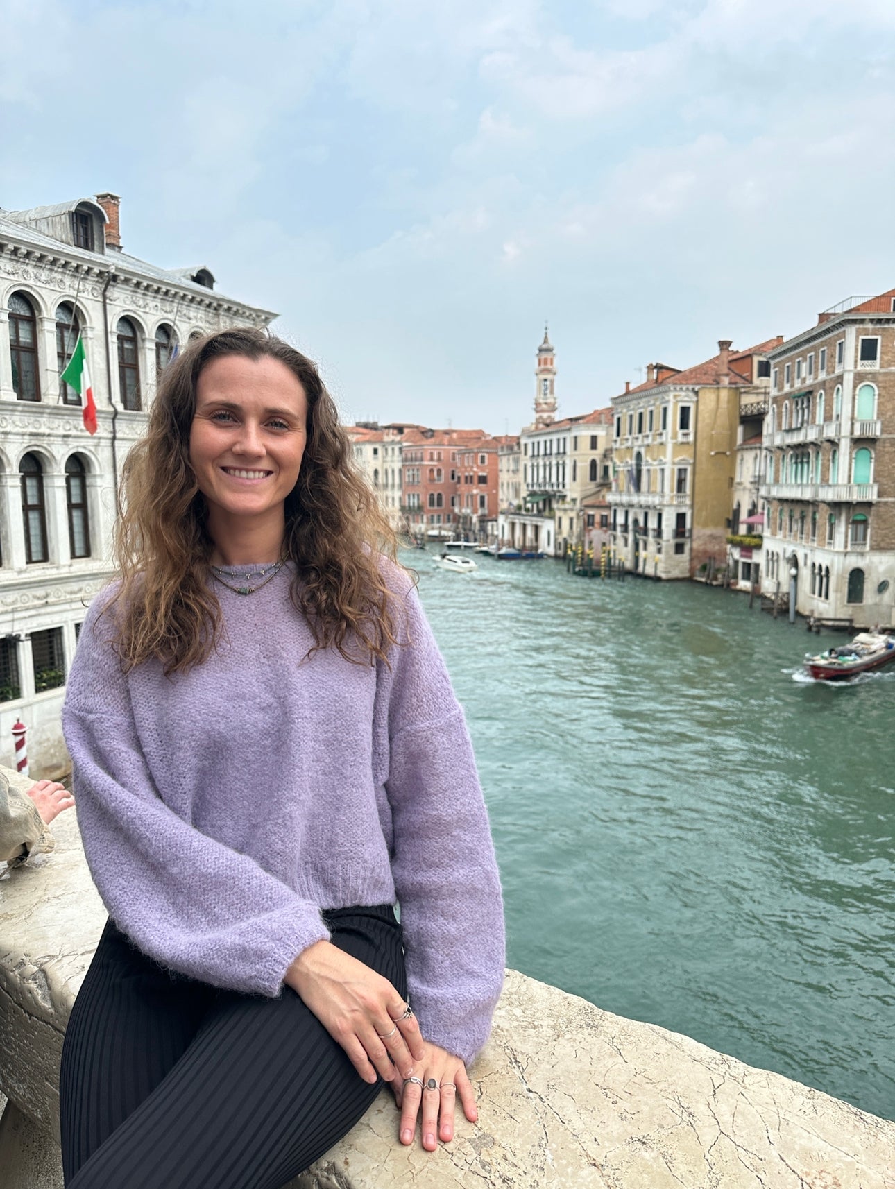 Woman in lavender sweater sitting by a Venetian canal, historic buildings in background