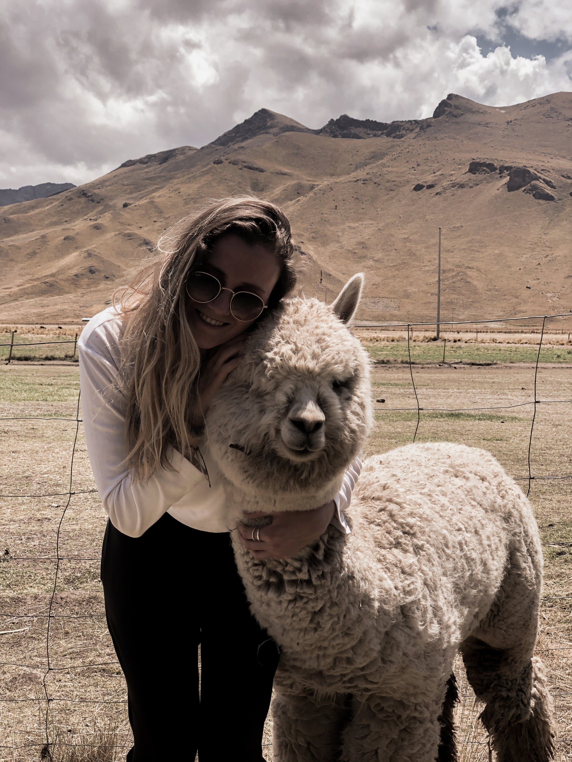 Woman hugging alpaca in grassy mountain landscape under cloudy sky
