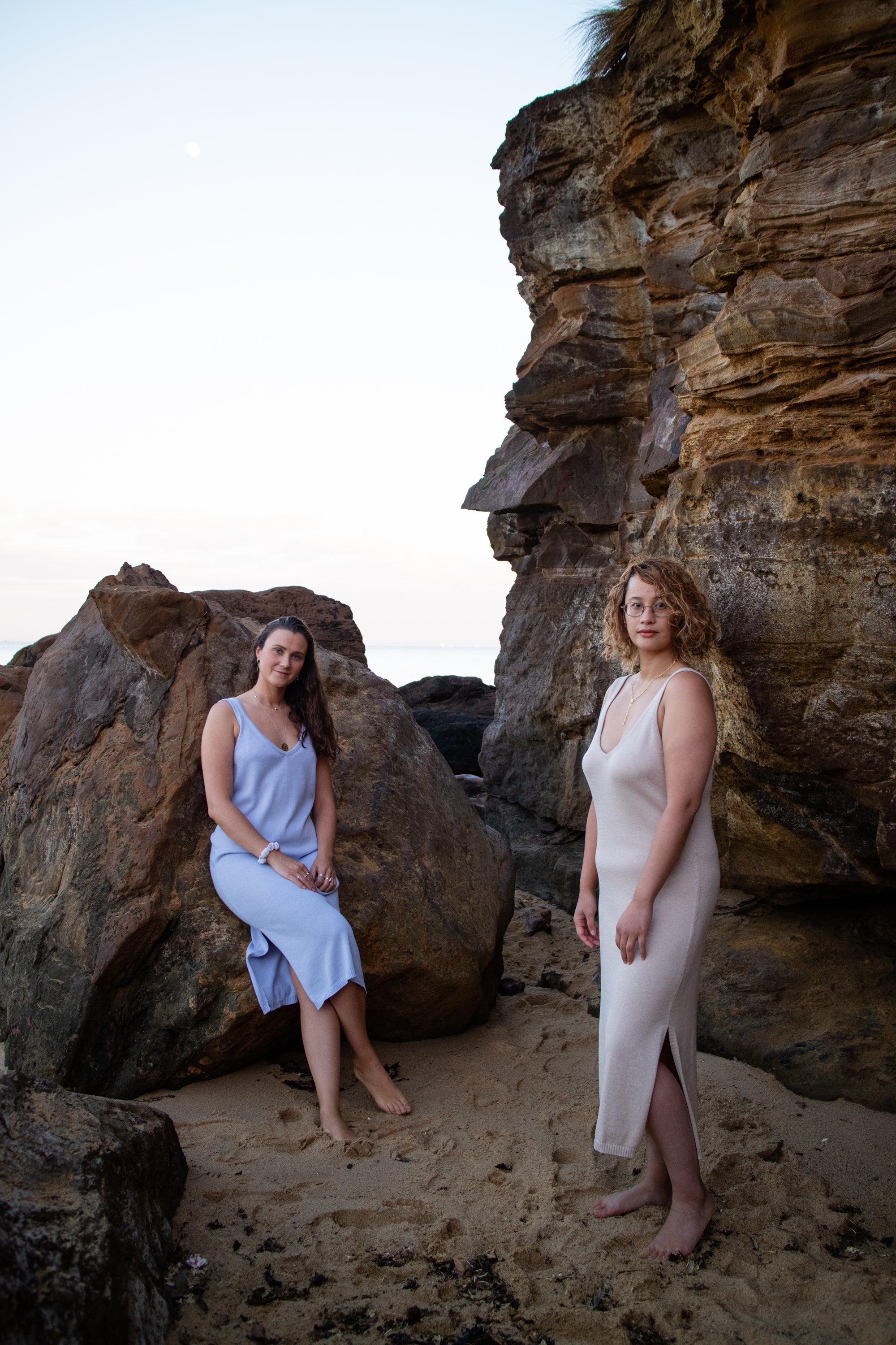 Two women in minimalist dresses pose on a rocky beach at sunset