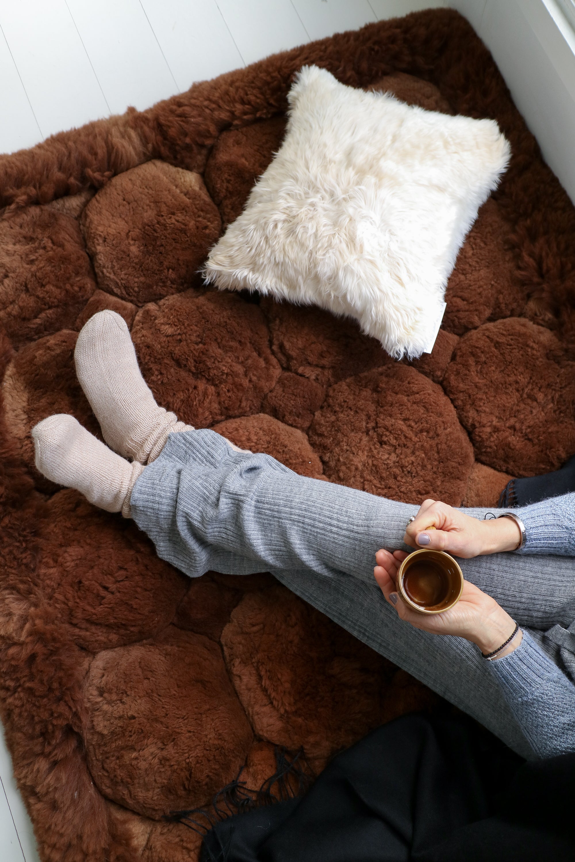 Person in cozy socks and knit pants relaxing on brown sheepskin rug with fluffy pillow and coffee