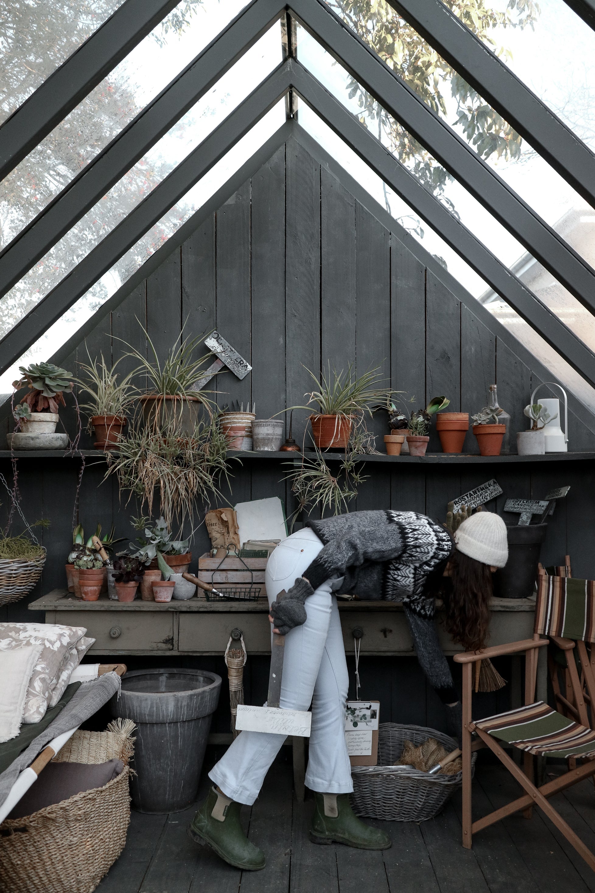 Woman in sweater and beanie tending plants in cozy greenhouse with potted greenery