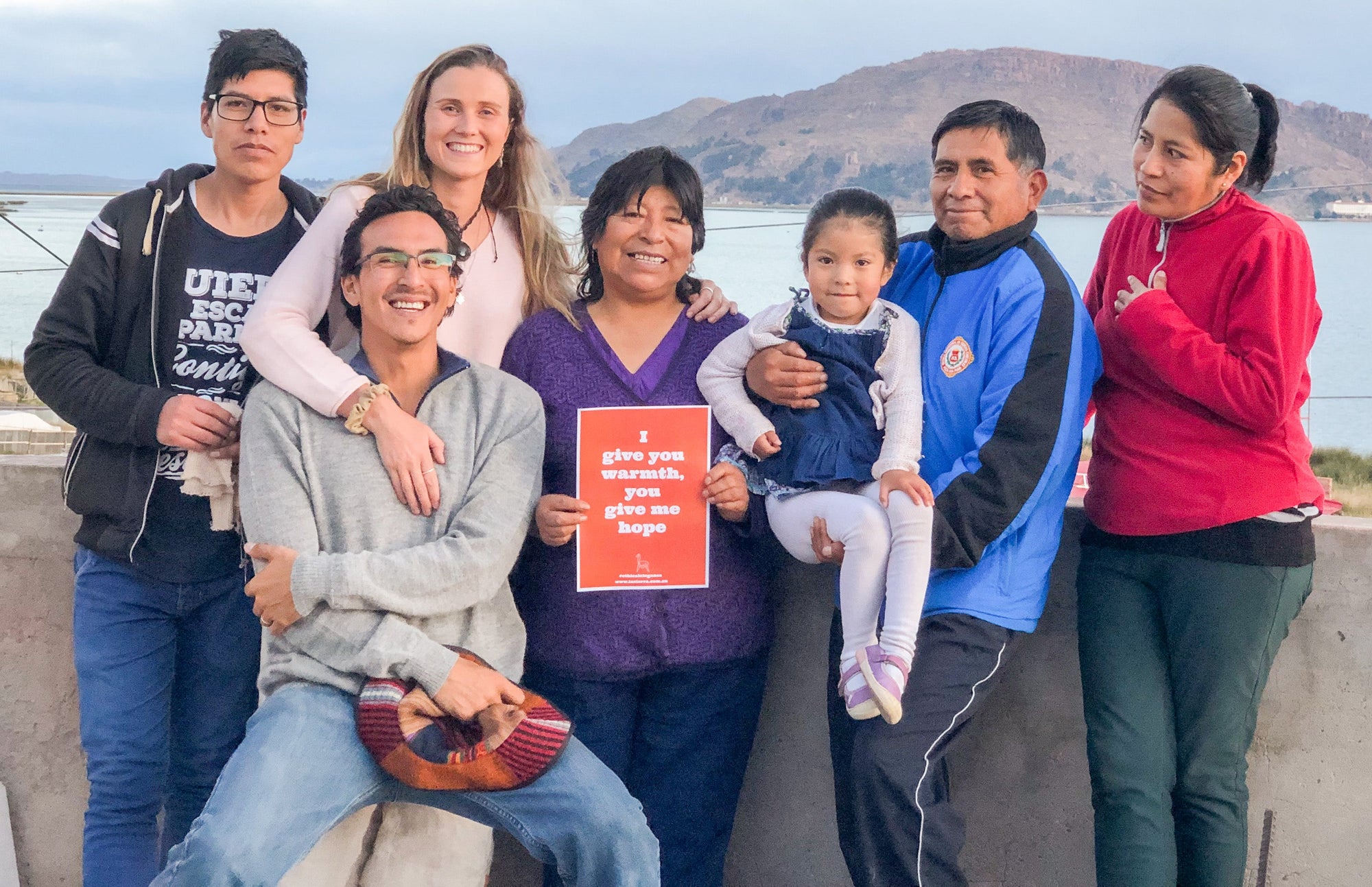 Smiling diverse group standing outdoors by a lake, woman holding a sign, mountain background