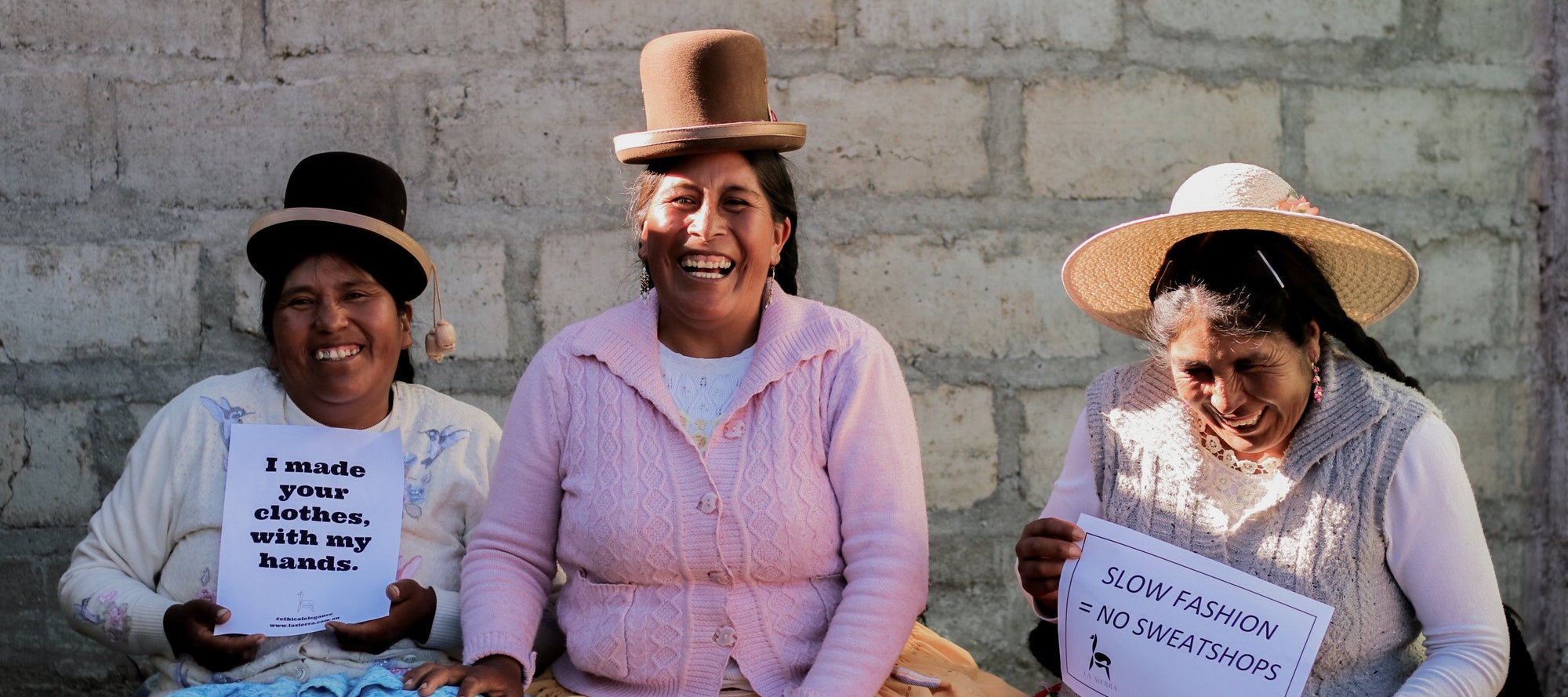 Three women artisans smiling, holding signs promoting ethical handmade fashion, against a brick wall.