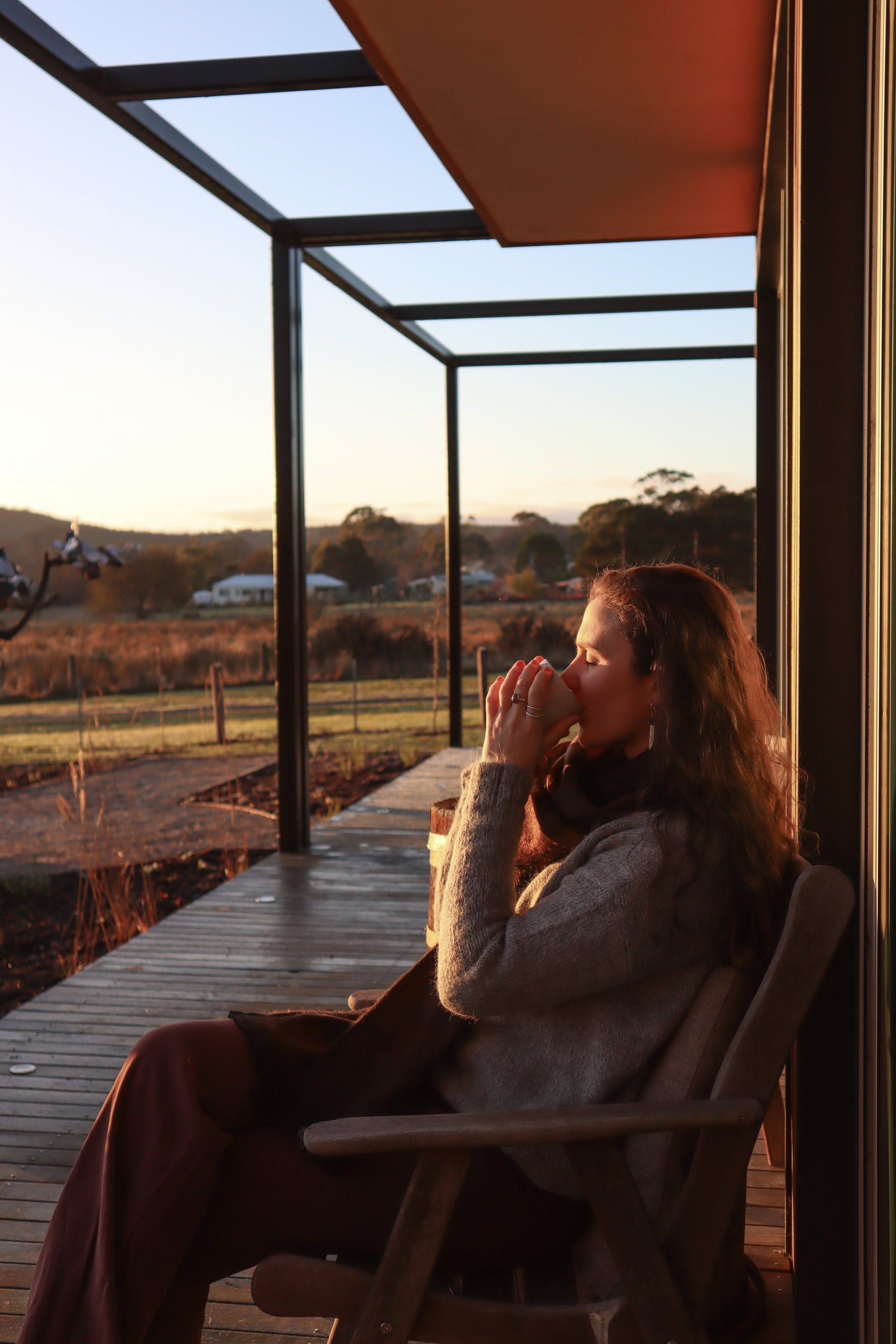 Woman in cozy sweater enjoying a warm drink on a porch at sunset, countryside view