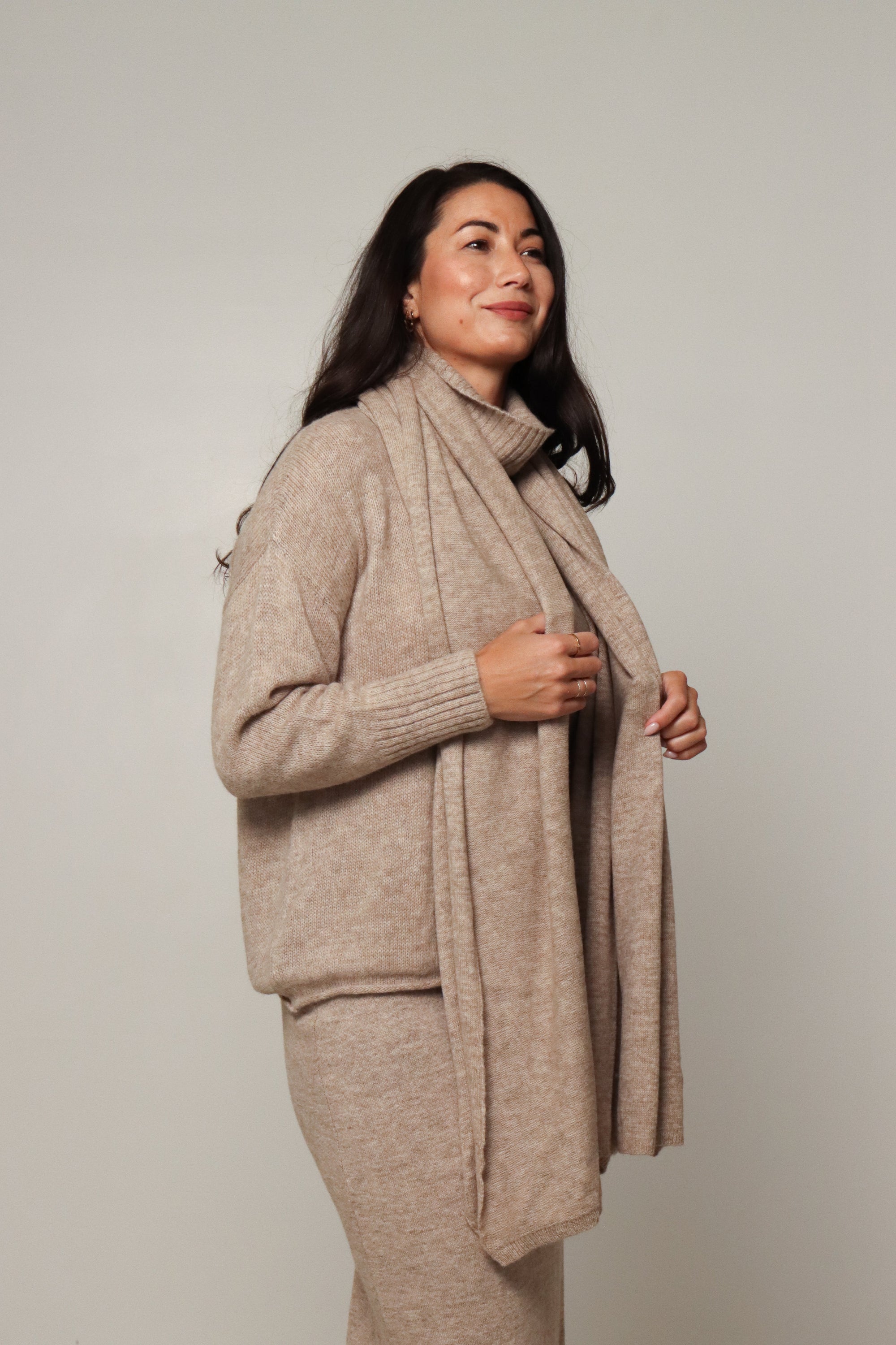 Woman wearing beige alpaca wool sweater, matching scarf and pants, neutral background
