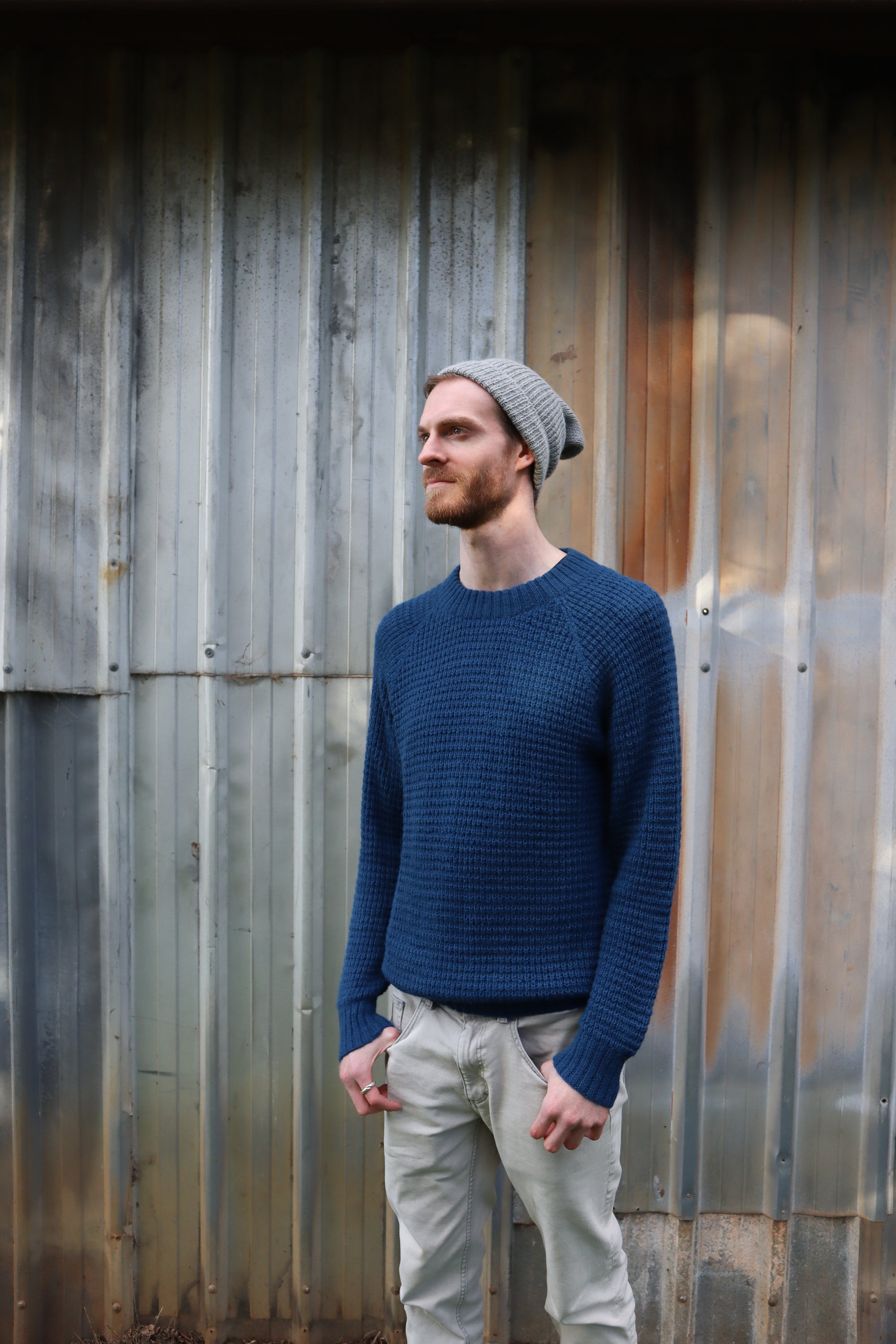Man wearing a blue knit sweater and gray beanie against a weathered metal wall