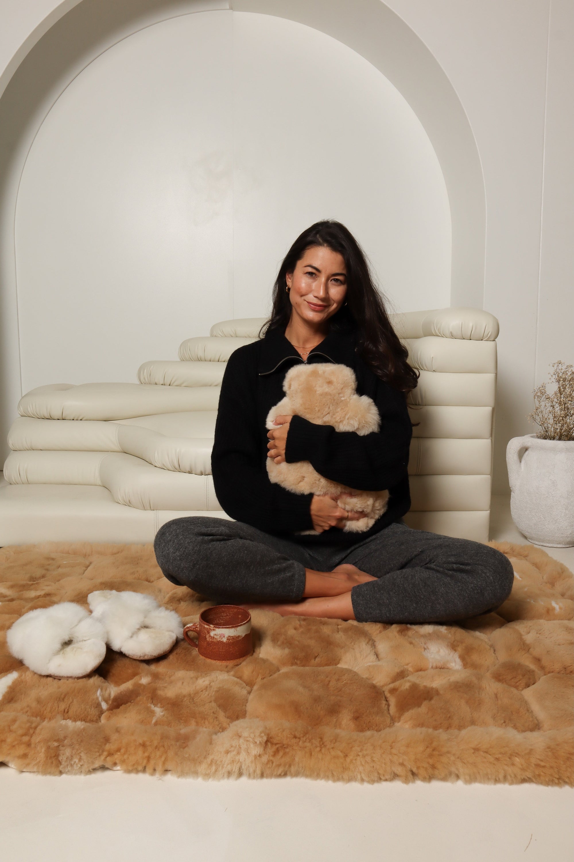 Smiling woman relaxing on cozy fur rug with slippers, blanket, and mug for Mother's Day gift guide