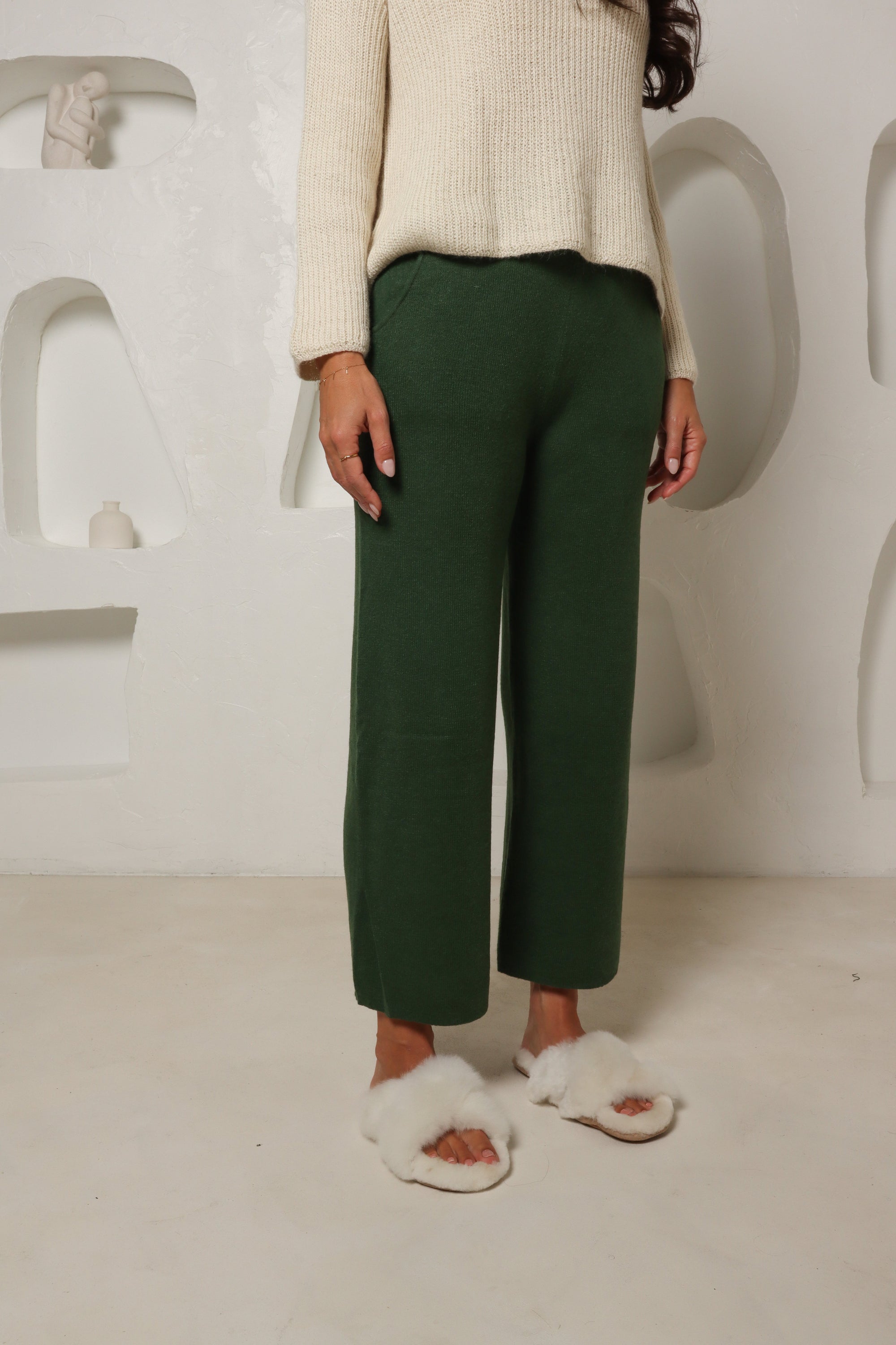 Woman wearing green alpaca pants, cream sweater, and fluffy slippers in a modern room